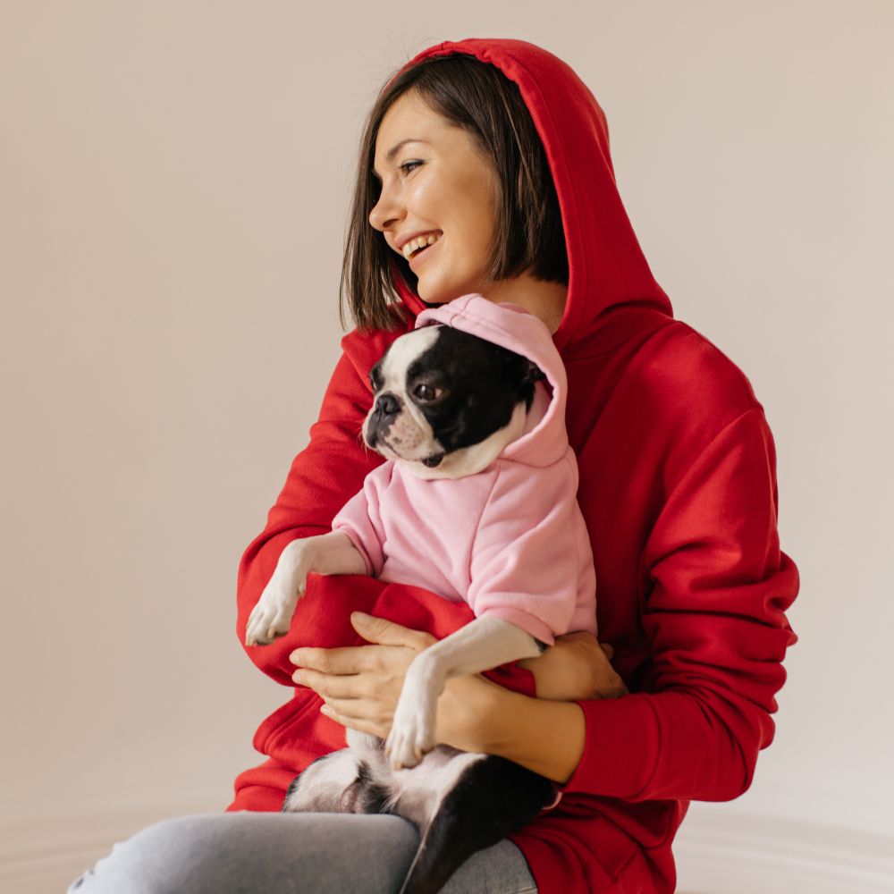 Woman wearing a red hoodie holding a dog in a pink hoodie against a beige background