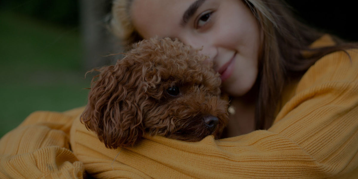 A woman in a mustard yellow ribbed sweater hugging a small curly-haired brown dog outdoors.